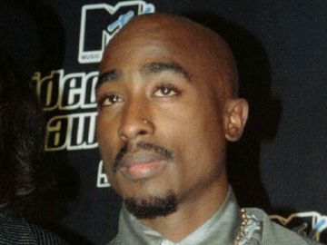 En mann med svart skjegg og svart bart og uten hår ser alvorlig ut og står foran en svart bakgrunn.