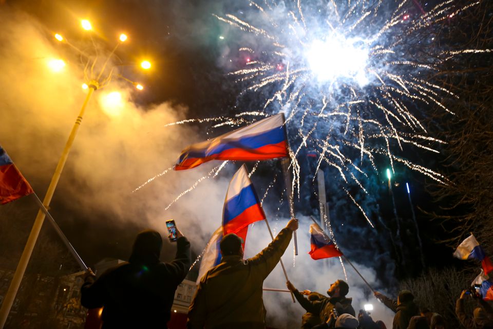 Et bilde tatt utendørs i mørket viser røyk, gatelys og fyrverkeri som eksploderer, mens folk veiver med russiske (hvite, blå og røde) flagg.