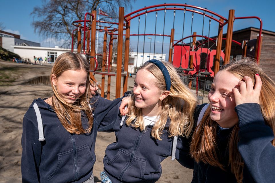 Tre jenter som står med armene rundt hverandre og smiler foran et lekestativ