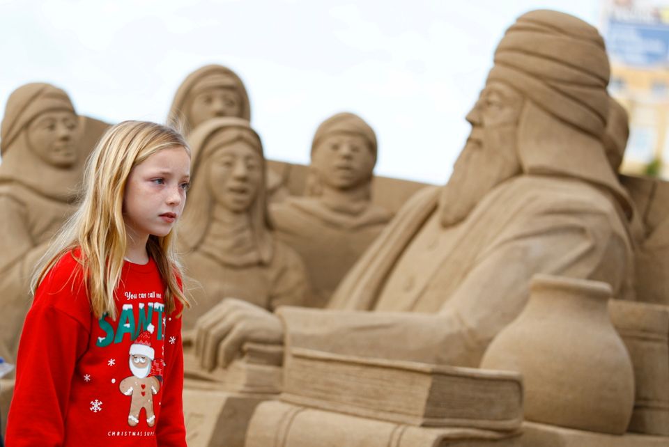 En jente med langt, blondt hår og rød genser med julemotiv på stirrer skeptisk på enorme menneskelige sandskulpturer.