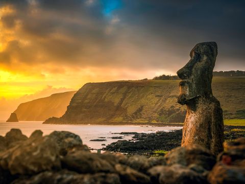 Én høy stein-høvding står ved strandkanten mens himmelen er skydekket og gløder gult under solnedgangen.