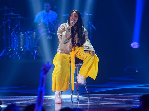 Ung kvinne med gul bukse og grå jakke sitter på en barkrakk på en scene og synger i en mikrofon