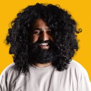 Tett portrett av en smilende mann med svart, krøllete hår og skjegg mot en knallgul bakgrunn.