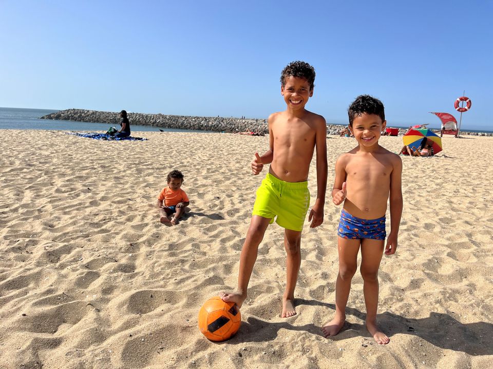 To gutter i badebukse står og smiler ved en fotball på en strand, mens en veldig liten gutt sitter i sanden bak dem.