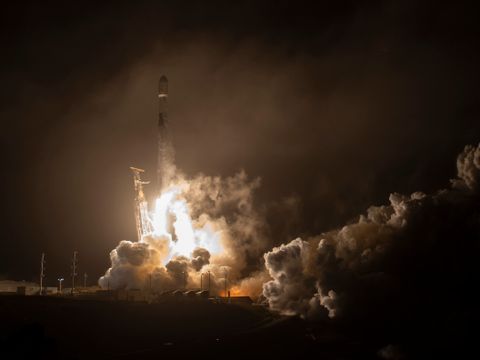 Bildet er tatt fra bakkenivå og vinklet opp mot en mørk nattehimmel, mens en rakett skyter oppover i luften og tilbakelegger masse røyk. 