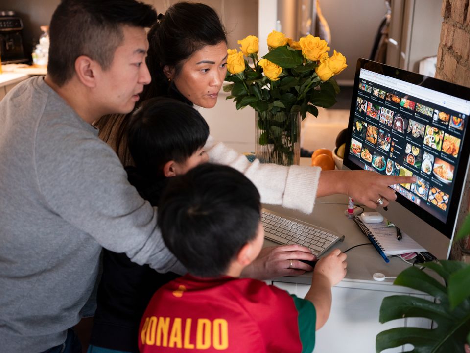 En mamma, pappa og to små gutter står samlet ved en stor PC-skjerm med bilder av mange matretter på og mammaen peker på noe på skjermen. 
