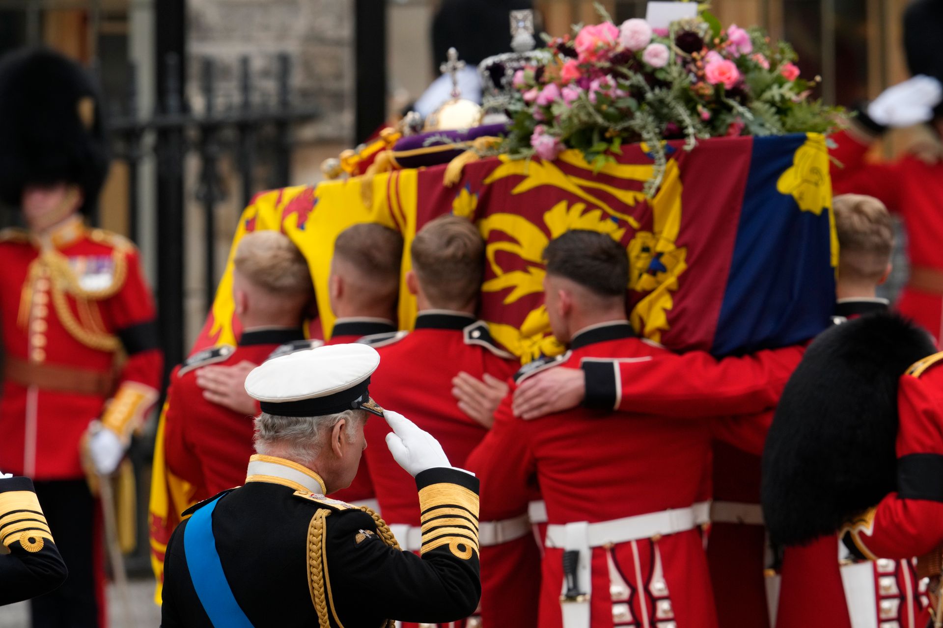 Åtte menn med røde jakker bærer en kiste som er dekket av et flagg med et slags symbol på, i rødt, gult og blått, mens en mann i forgrunnen hilser mot kisten, og oppå kisten ligger det blomster og en krone. 