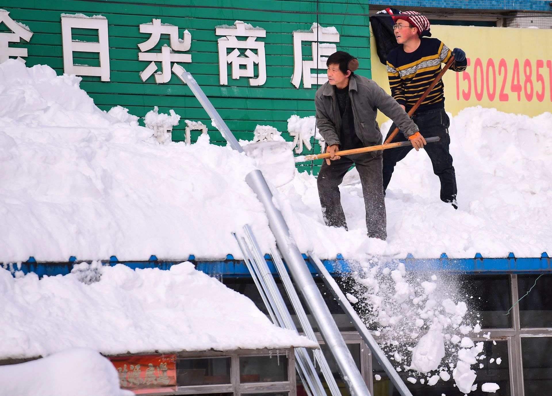 To menn står oppå et tak og bruker spader for å fjerne store mengder med snø.