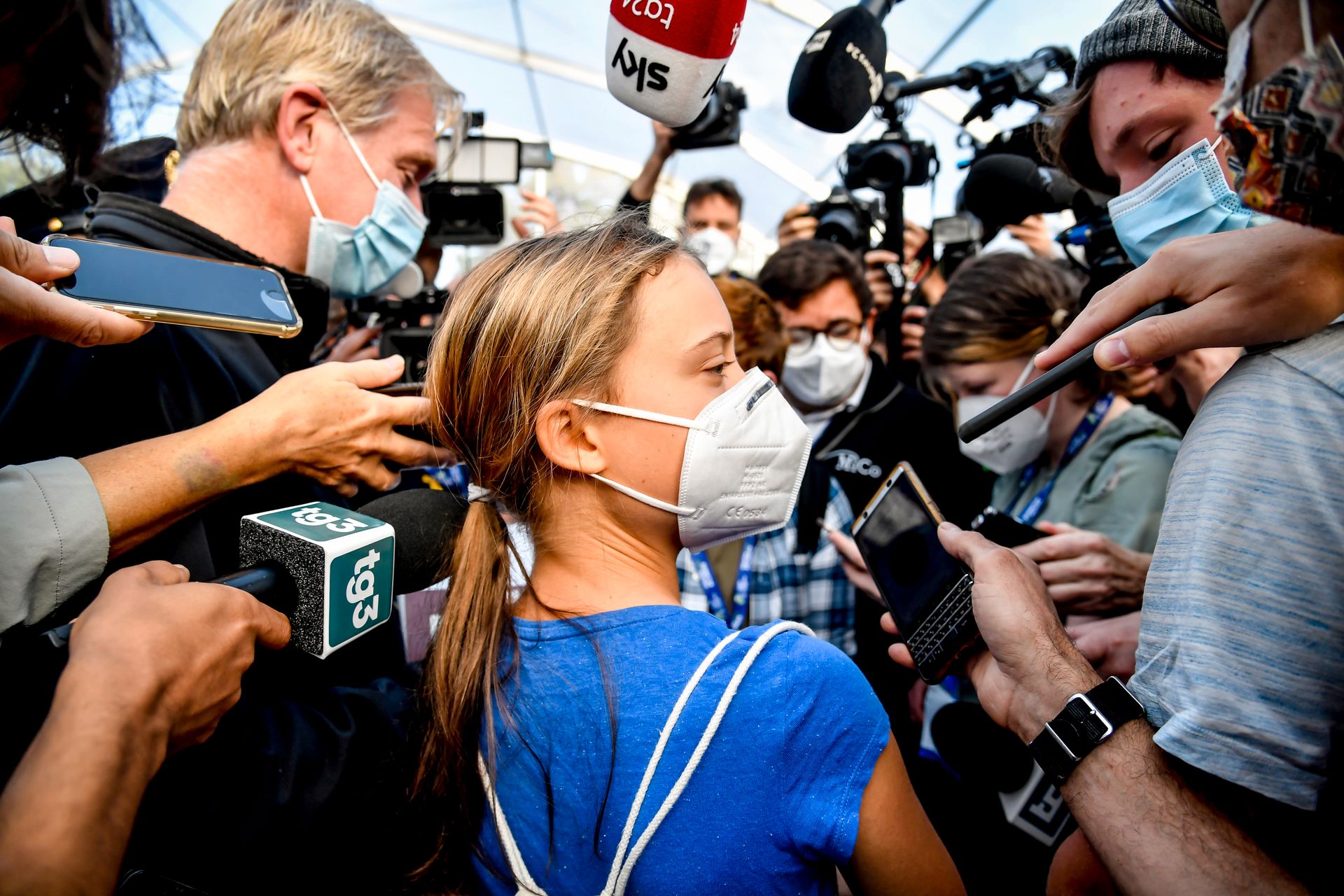 Et portrett tett på Greta Thunberg, med ansiktsmaske på, omgitt av mange reportere med mikrofoner og kameraer.