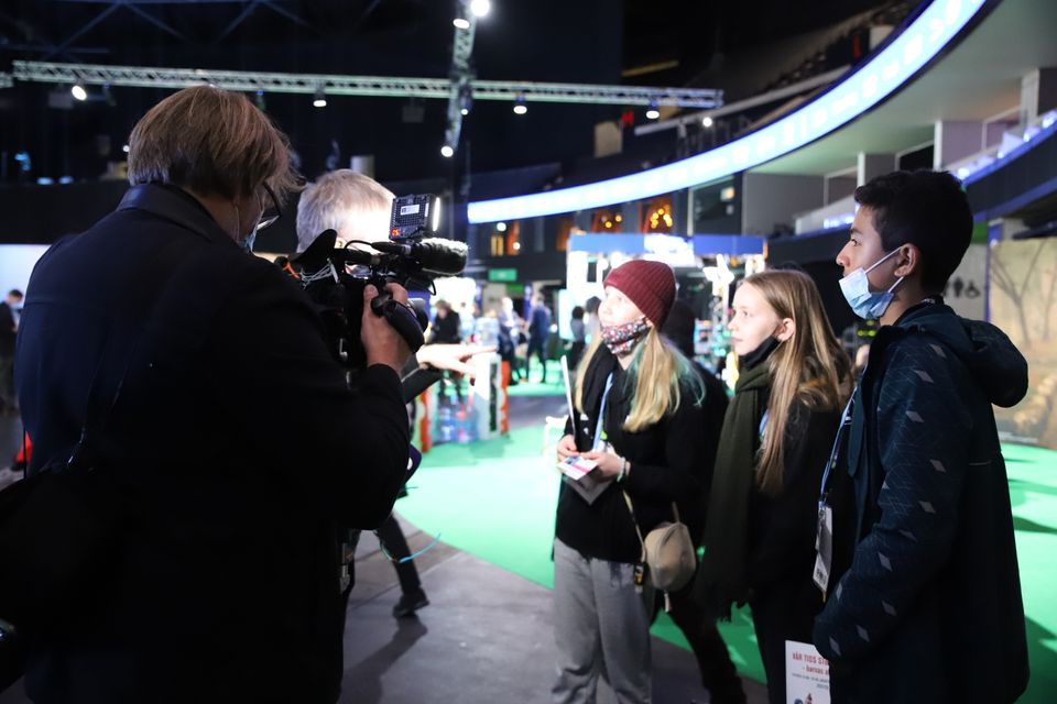 To jenter og en gutt står og blir intervjuet av en journalist og en mann med et stort filmkamera filmer dem. 