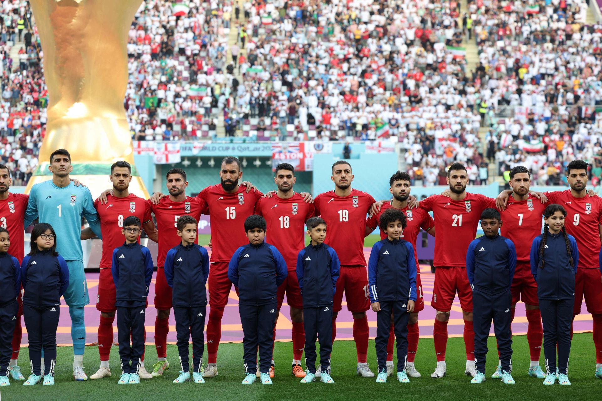 Mange spillere i røde drakter og en mindre gutter med blå drakter står på rekke på en fotballbane, med armene på hverandres skuldre, foran en full tribune.