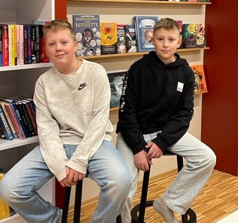 To unge gutter sitter på hver sin stol med hyller med bøker i bakgrunnen og begge smiler.