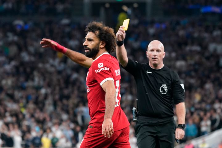 En fotballspiller med rød drakt og svart, krøllete hår er irritert fordi han får et gult kort av dommeren.
