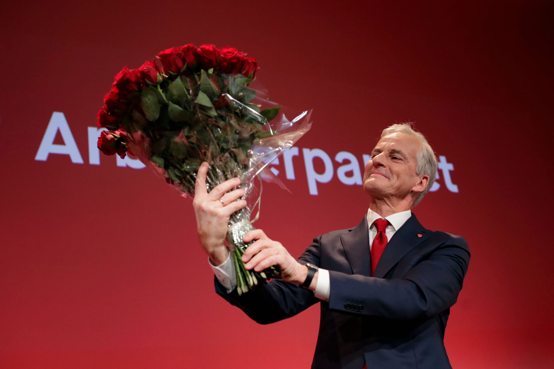 Støre ses nedenifra, mens han står foran en rød vegg med Aps logo, på scenen under Aps valgvake, og ser på den store rosebuketten han løfter opp foran seg.