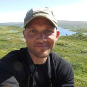 En mann med grå caps som tar en selfie ute i et grønt naturområde. 