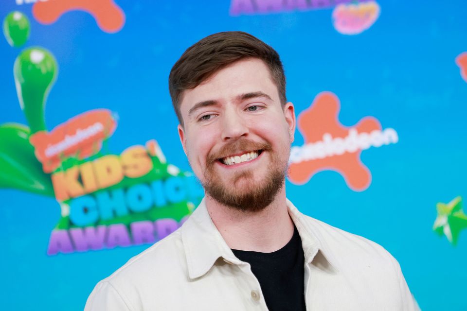 Youtuberen MrBeast, med kort brunt hår, smiler og står foran en fargerik plakat.