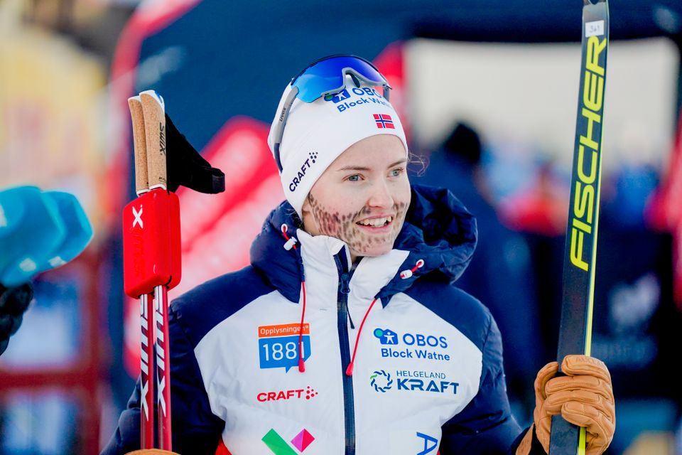 En kvinne med hvit jakke og hvit lue står med ski og staver i hendene og har farget mange svarte prikker i ansiktet som ser ut som skjegg.