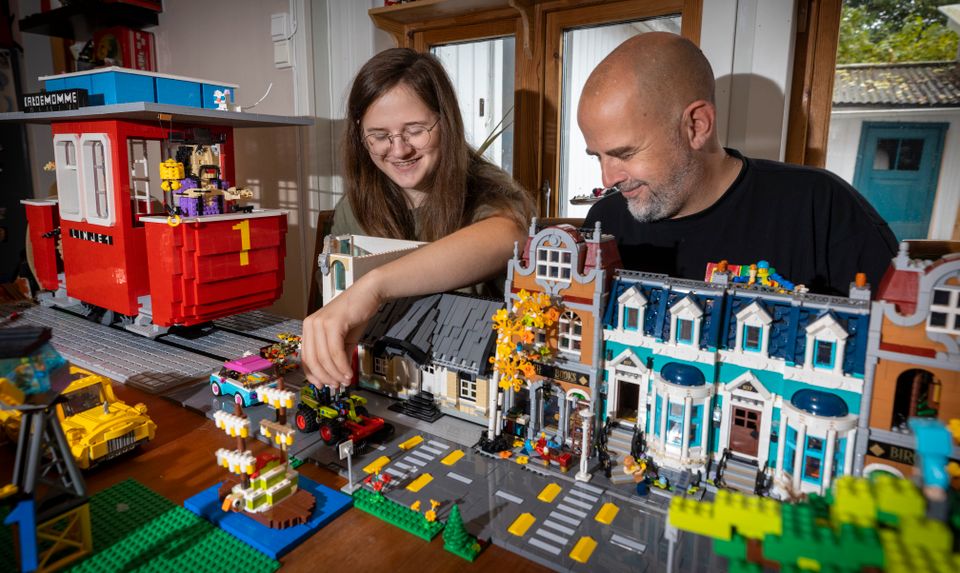 En gutt med briller og langt hår og en mann uten hår sitter bak og ser ned på en by bygget av lego med hus og veier som står på et bord. 