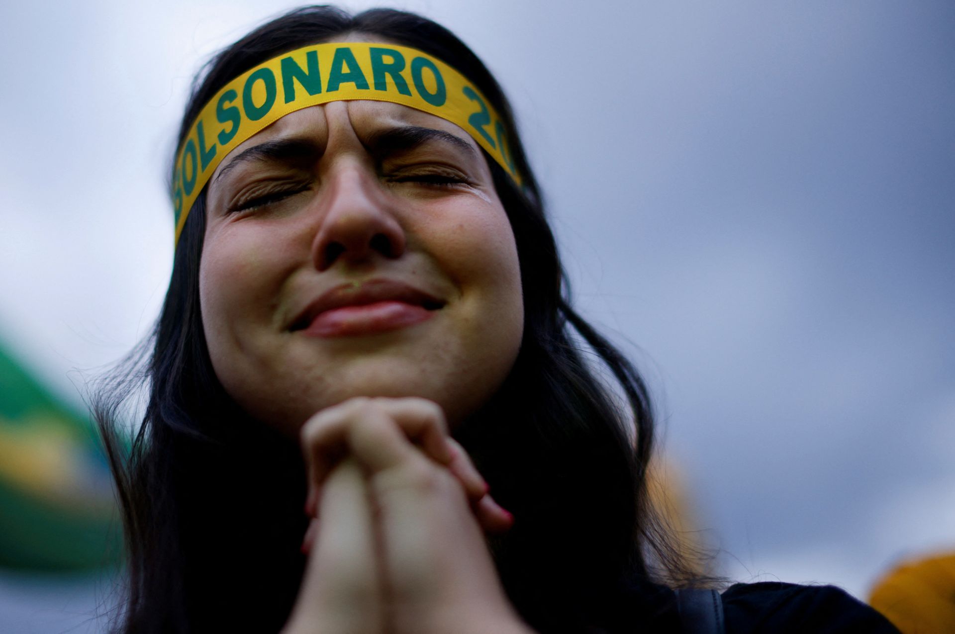 Kvinne folder hendene mens hun lukker øynene. Det ser ut som om hun ber. Kvinnen har på seg et Bolsonaro-bånd rundt hodet.