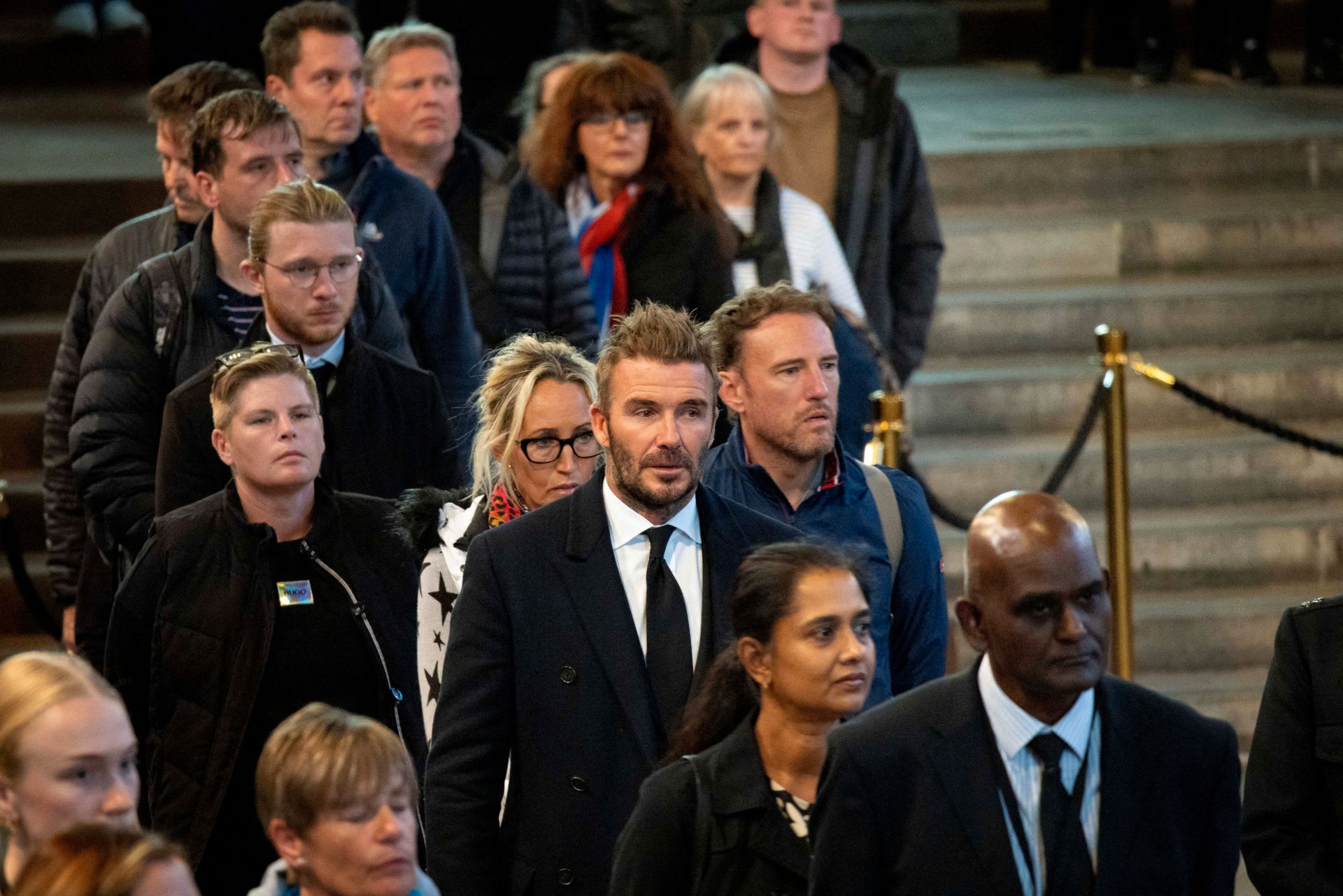 Mange mennesker står i kø i en steintrapp, og midt i bildet står en mann med mørk dress, lyst hår og litt skjegg: David Beckham. 