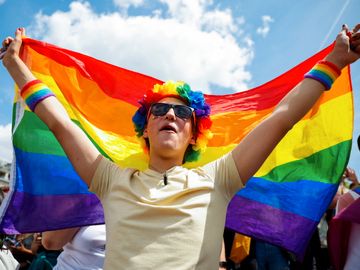 En person med fargerik parykk og gul T-skjorte, holder et regnbue-flagg bak seg.