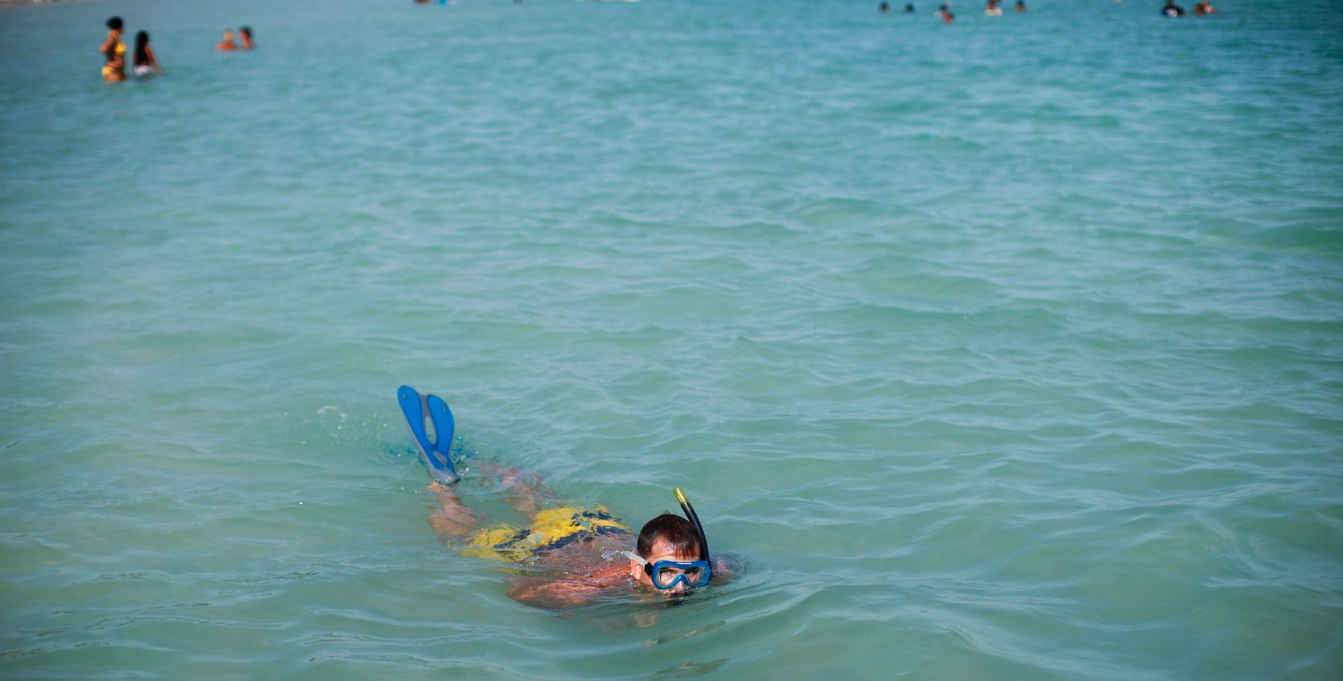 En mann med badeshorts, snorkel og svømmeføtter svømmer i turkist, rolig vann mens han kikker opp på fotografen.