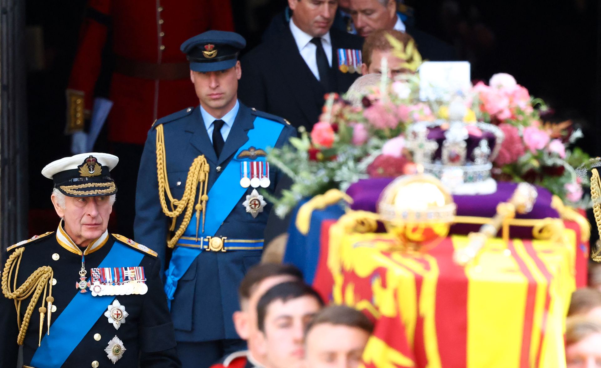 To menn, én gammel og en yngre, med hatter og blå jakker med utsmykkinger og blå bånd på, går bak en kiste som er dekket av blomster, et flagg og har en stor krone oppå.