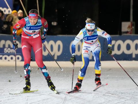 En kvinne med mørkt hår og rød skidrakt sprinter om kapp med en kvinne med hvit, blå og gul drakt på ski.