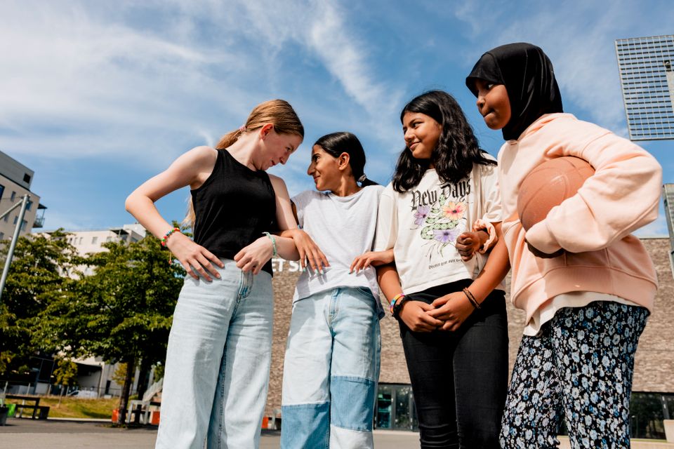 Fire jenter, en med langt mellomblondt hår, to med langt svart hår og en jente med hijab og en basketball under armen, holder hverandre rundt albueleddet og smiler mot hverandre. 
