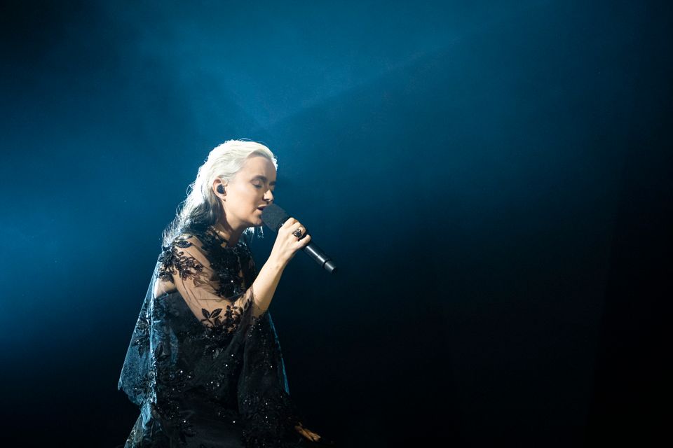 En ung kvinne med langt lyst hår og sort kjole som står på en scene og synger inn i en mikrofon. 