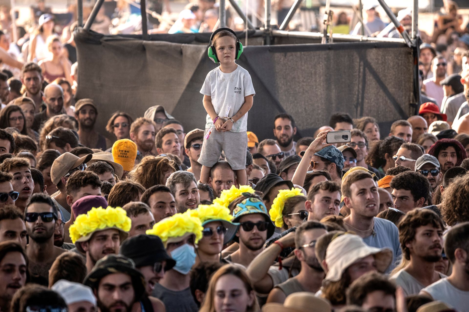 En gutt med hvit T-skjorte, grå shorts og grønne hørselsvern, står på skulderen til en mann i en stor folkemengde.