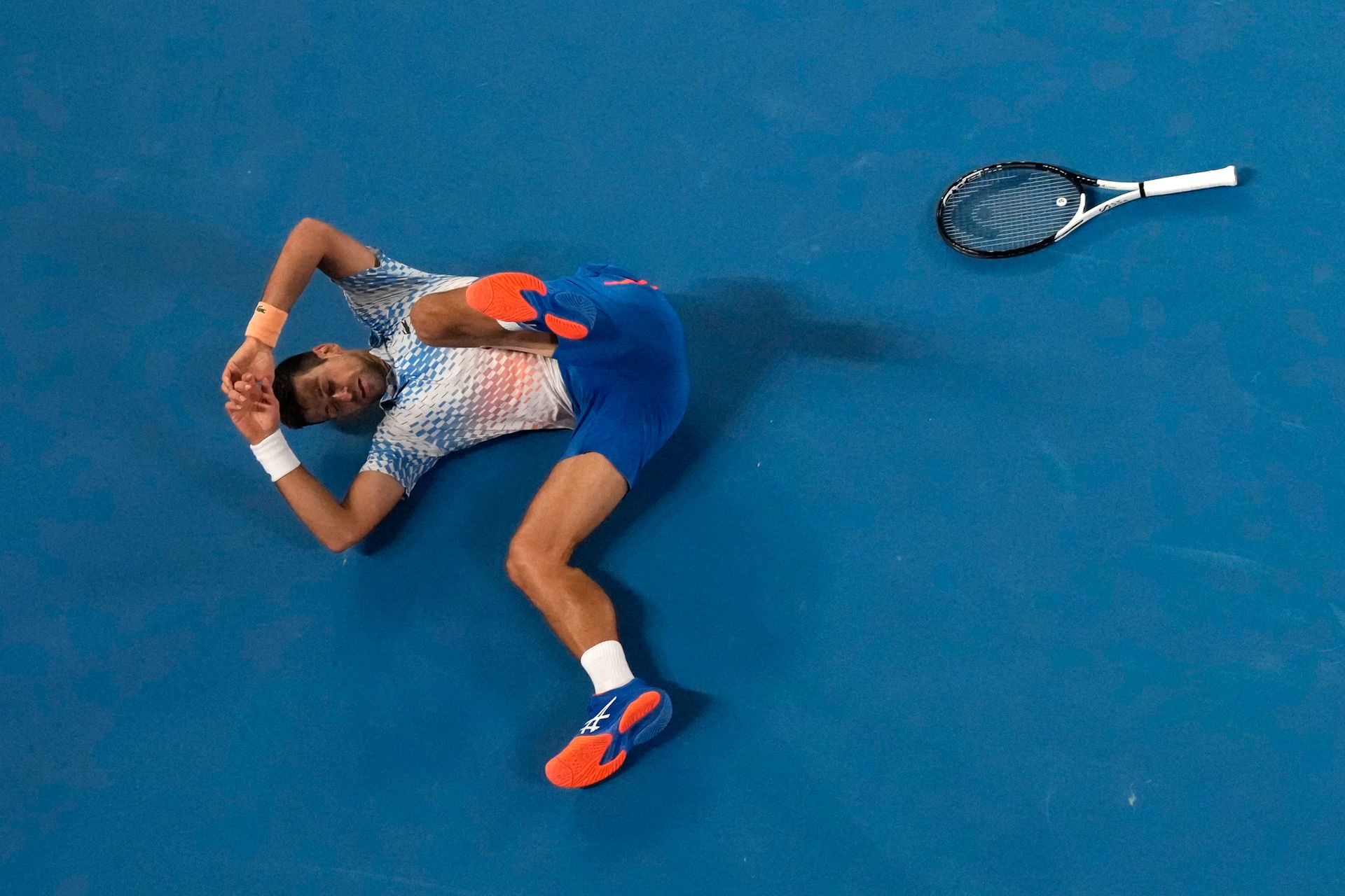 En mann med blå shorts og røde sko ligger på bakken, med beina i været og hendene over hodet, ved siden av en tennis-racket.