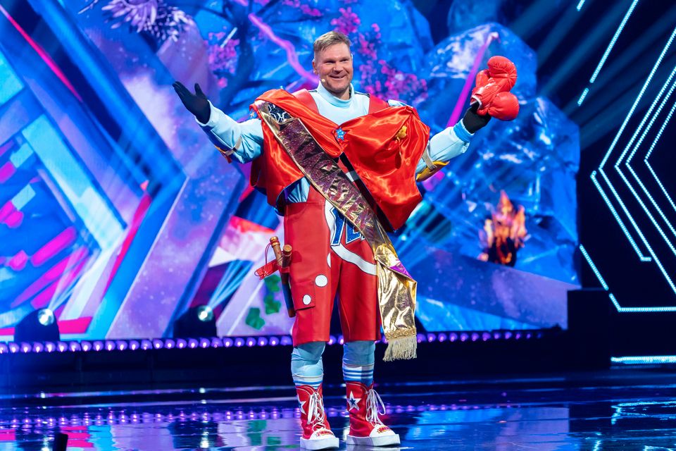 En mann står på en scene og smiler og har på seg et stort blått kostyme med rød drakt