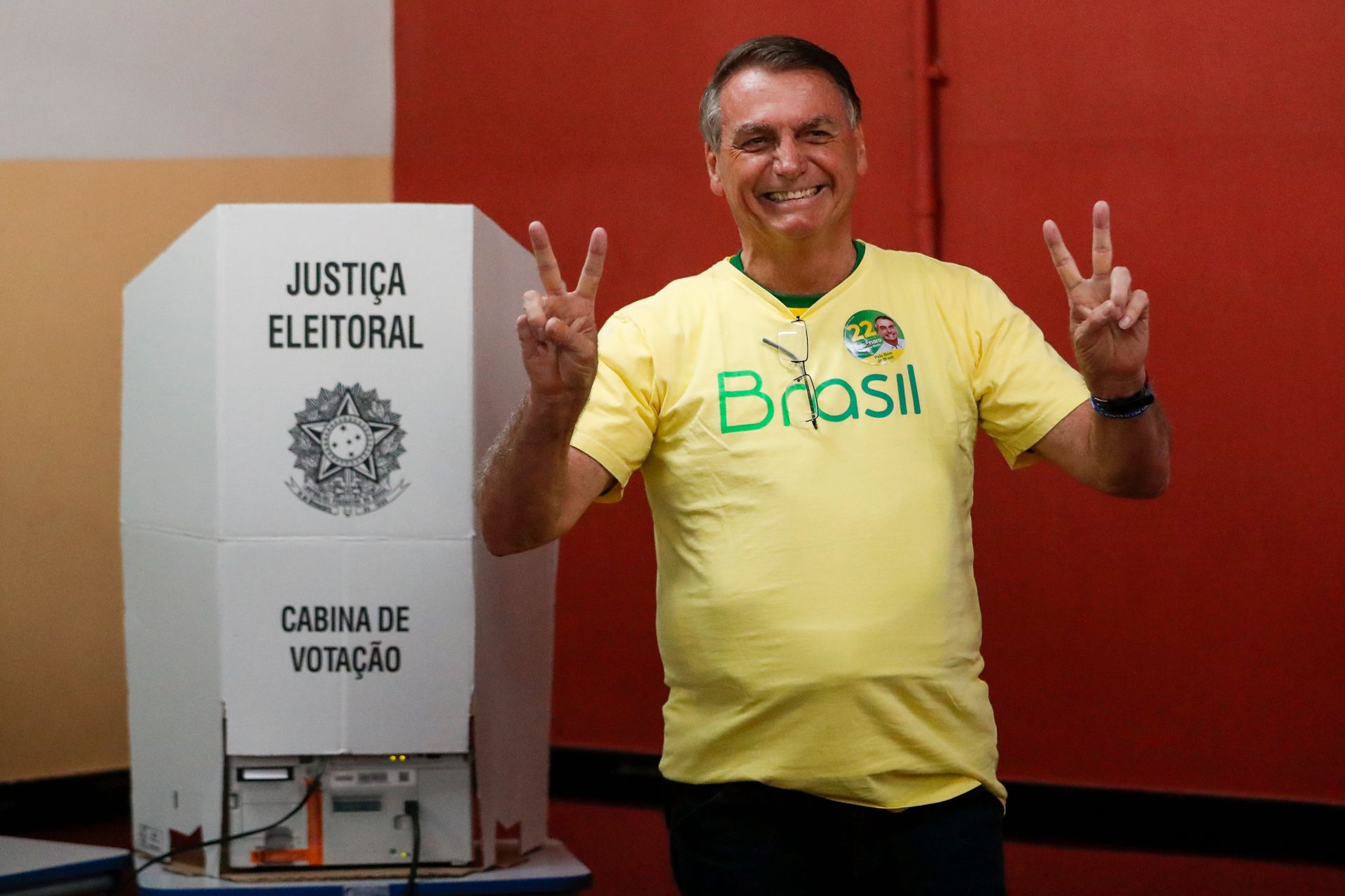 Bolsonaro i en gul t-skjorte viser peace-tegn med begge hendene i et stemmelokale