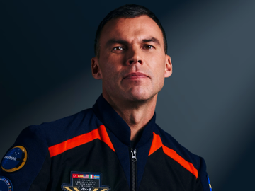 En mann med astronaut-uniform smiler lett mot fotografen fra et rom med mørk bakgrunn.