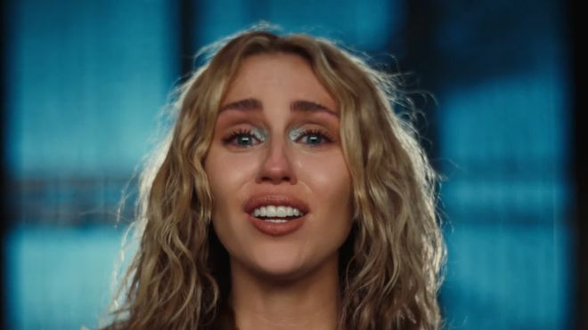 Nærbilde av Miley Cyrus, en kvinne med bølgete, blondt hår og Mikke Mus-T-skjorte, som synger trist inn i kameraet.