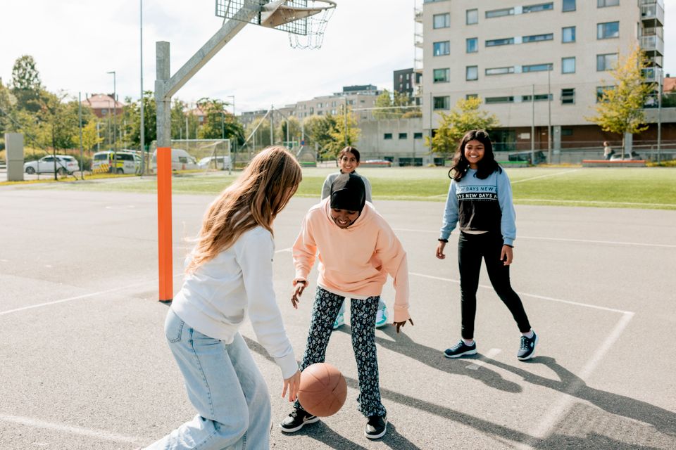 Fire jenter leker med en basketball like ved en kurv, på en asfaltbane på sommertid.