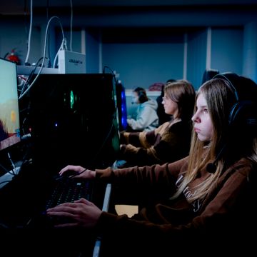 En jente med langt brunt hår sitter konsentrert foran en lysende PC og spiller. 