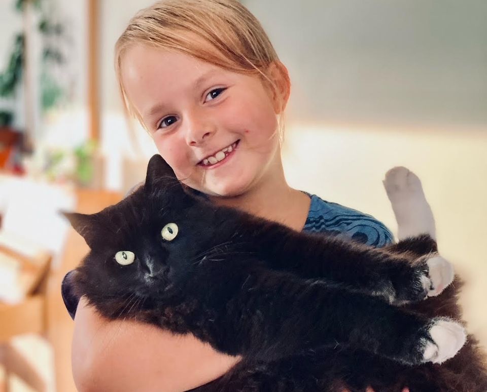 En ung jente med blondt hår holder en svart katt i armene og smiler.
