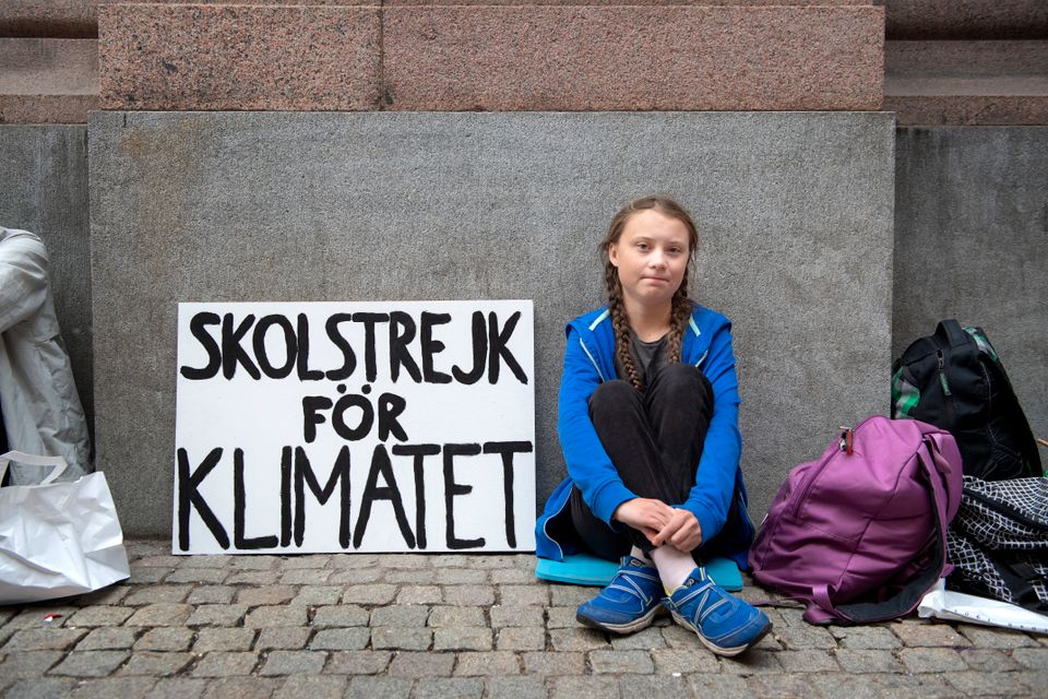 En jente med langt, mørkeblondt hår og blå genser sitter foran et skilt hvor det står skolestreik for klima