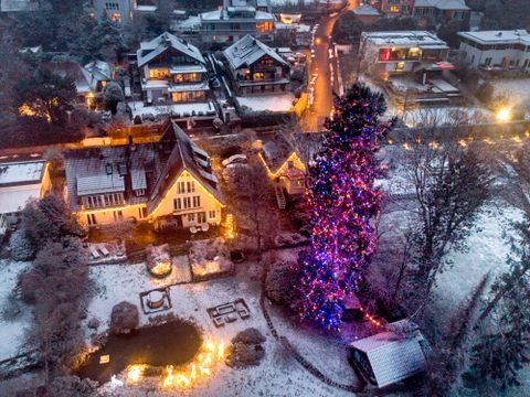 En dronesilde viser et lite område i en landsby der hus er opplyst av gylne julelys og det står et enormt grantre med rosa og lilla lys på i en hage.