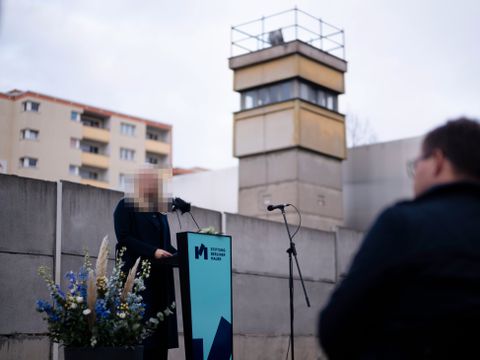 En kvinne med lang kåpe og sensurert ansikt står og taler foran en folkemengde ved restene av Berlinmuren.