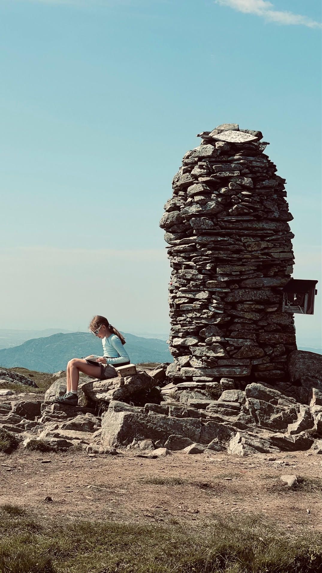 Jente med langt hår og blå genser sitter ved en stor varde og skriver i gjesteboka for fjelltoppen.