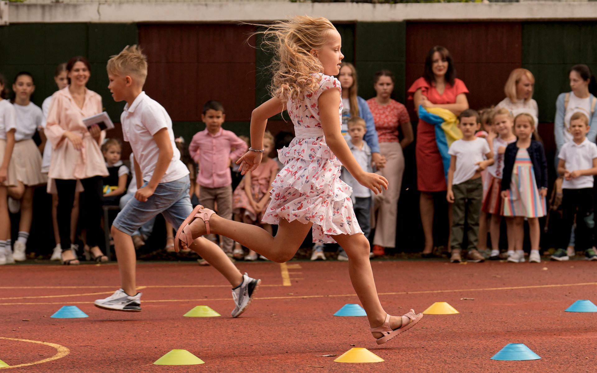 En lyshåret gutt og jente løper smilende i motsatte retninger over rød grus med fargerike, små kjegler på bakken.