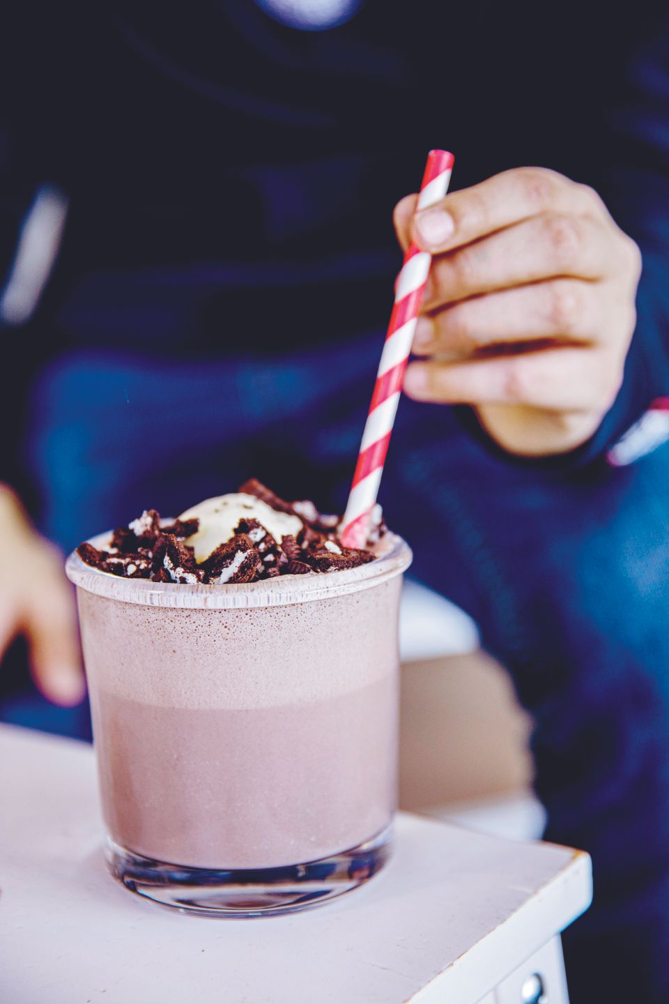 En hånd holder et rødt og hvit-stripet sugerør nedi et glass med sjokoladefarget drikk og Oreo-smuler på toppen.