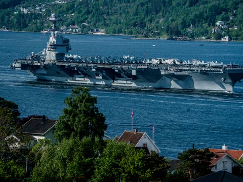 Et enormt, grått skip med flere titalls jagerfly på dekk, seiler inn Oslofjorden, forbi små trehus, som båten er høyere enn. 