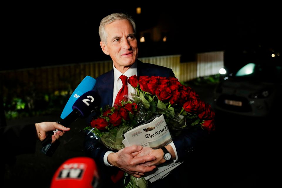 Jonas Gahr Støre står ute i mørket, med rødt slips og en bukett røde roser, mens reportere stikker mikrofonene sine mot ham.