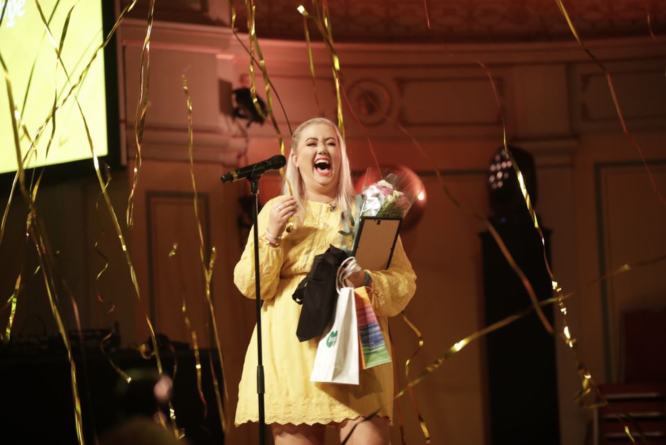 En ung kvinne med gul kjole står foran en mikrofon og er glad.
