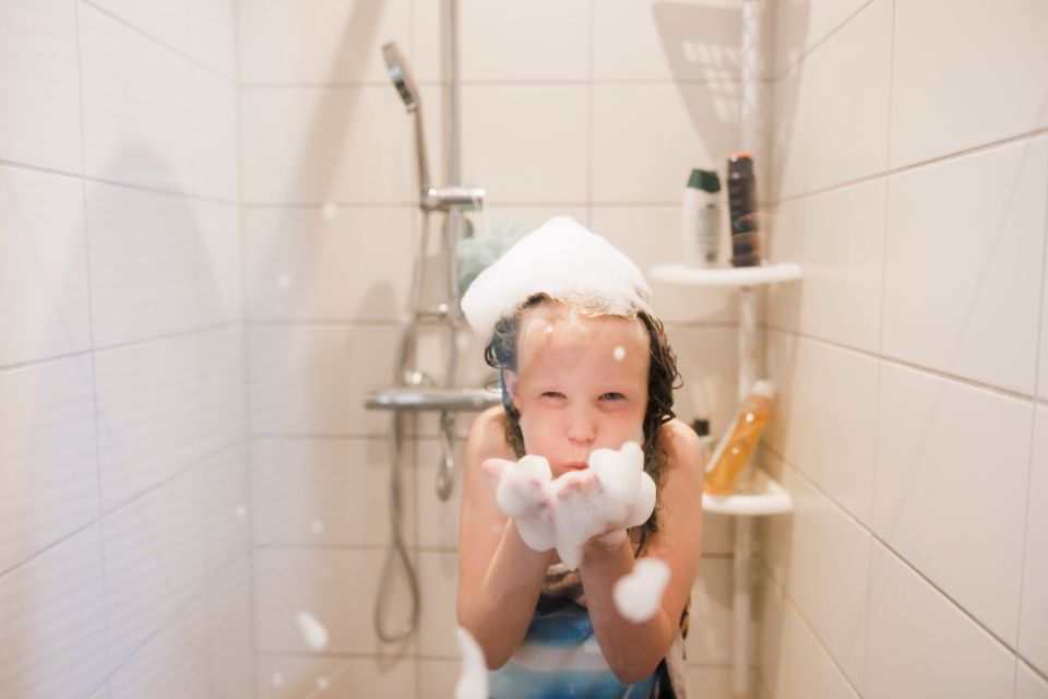 En jente står i dusjen med håndkle rundt kroppen og hvitt skum på hodet og blåser skum fra hendene.