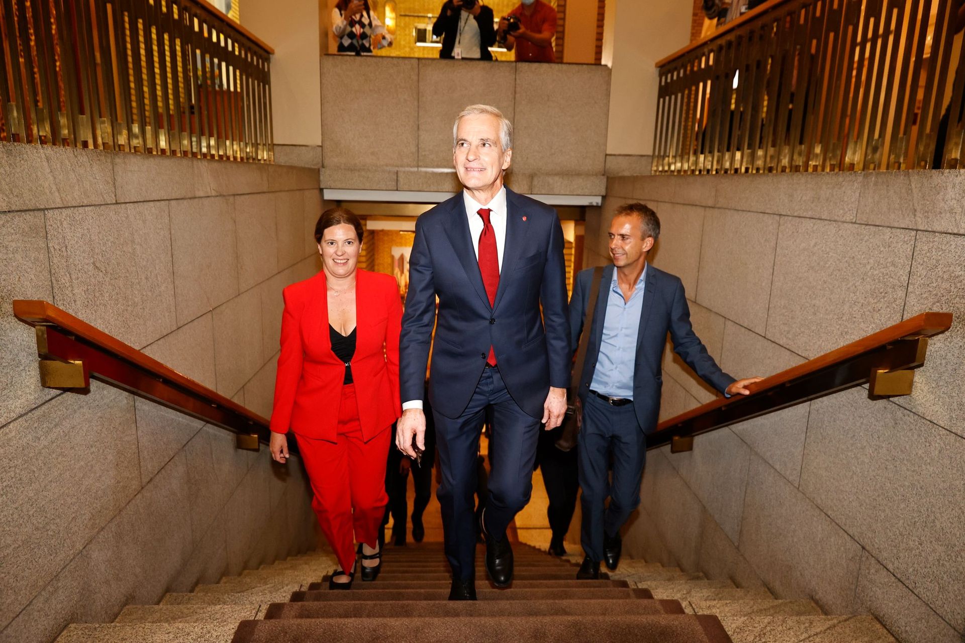 Jonas Gahr Støre går opp trappene inni Stortinget, ikledd dress og rødt slips, med medierådgiveren og en kvinne i rød dress like bak. 